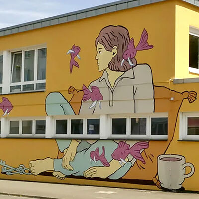Bild vergr&ouml;&szlig;ern: Im Juli 2024 malte die Künstlerin Greta von Richthofen dieses Kunstwerk innerhalb von einer Woche an die Fassade des Arbeiter‐Samariter‐Bunds in der Erzberger Straße. Die „Nachtwächterin“ wacht friedlich über das Schillerviertel. Gesellschaft leisten ihr dabei ihr Hund und einige fliegende Fische. Die fliegenden Fische tragen die Sorgen, Hoffnungen und Träume der Menschen in Notsituationen. Die idyllische Atmosphäre bildet auch einen Gegenpol zum Arbeitsalltag der Mitarbeitenden des ASB.