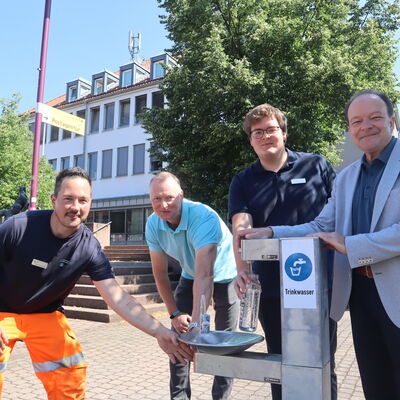 Bild vergr&ouml;&szlig;ern: Eine von drei Trinkwasserstationen steht in Oberkaufungen vor dem Rathaus: (v.r.) Bürgermeister Arnim Roß, Julius Krause (Klimaanpassungsmangager), Stefan Müller (Fachbereichsleiter Tiefbau, Stadt- und Landschaftsplanung, Infrastruktur) und Timo Windemuth (Gemeindebetriebe Kaufungen) stellten das neue Angebot vor.