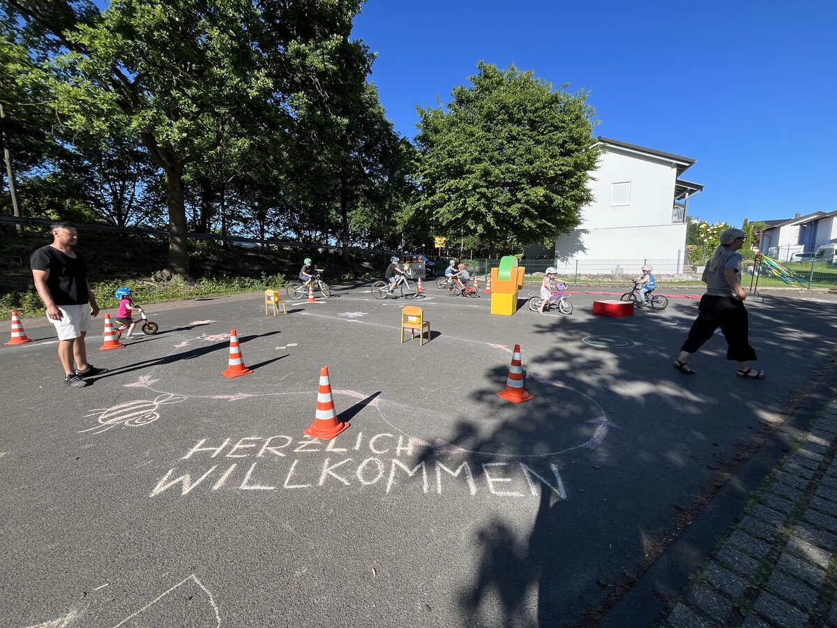 Auf einem Platz sind Verkehrshütchen und Stühle zu einem Parcours aufgebaut, es fahren Kinder auf Fahrrädern drum herum. Vorne im Bild auf dem Boden steht mit Kreide "Herzlich Willkommen".