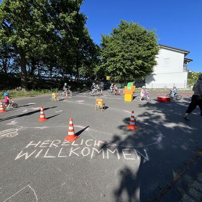 Bild vergr&ouml;&szlig;ern: Auf einem Platz sind Verkehrshütchen und Stühle zu einem Parcours aufgebaut, es fahren Kinder auf Fahrrädern drum herum. Vorne im Bild auf dem Boden steht mit Kreide "Herzlich Willkommen".